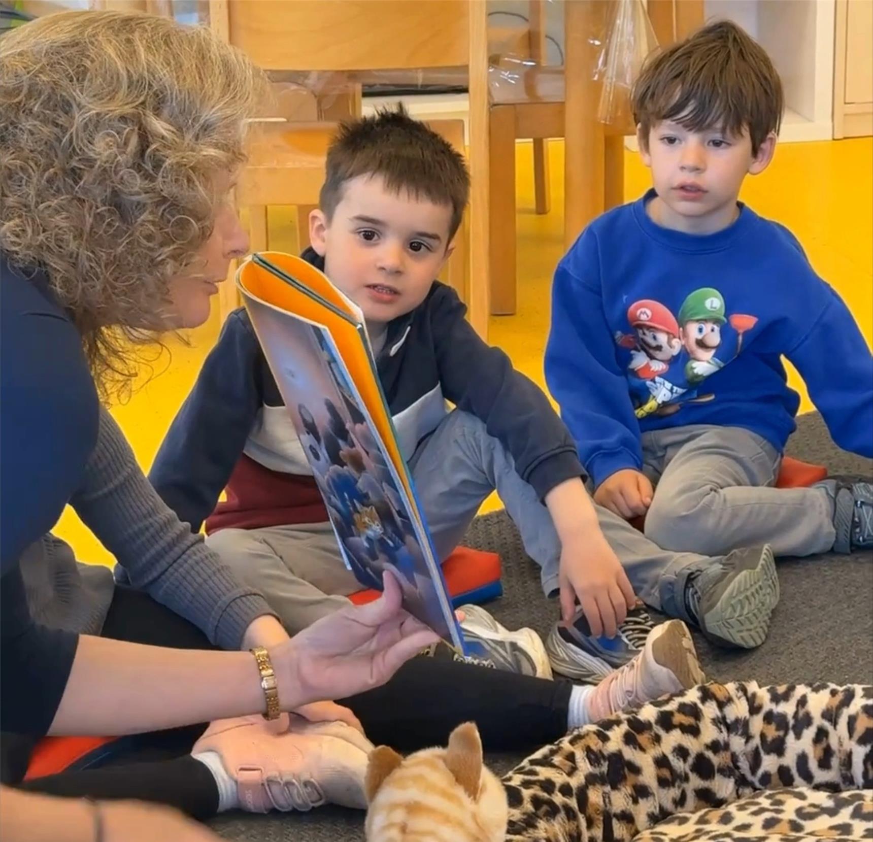 Zwei Kinder hocken auf dem Boden und blicken ganz gebannt auf eine Erzieherin, die ihnen ein Buch präsentiert.