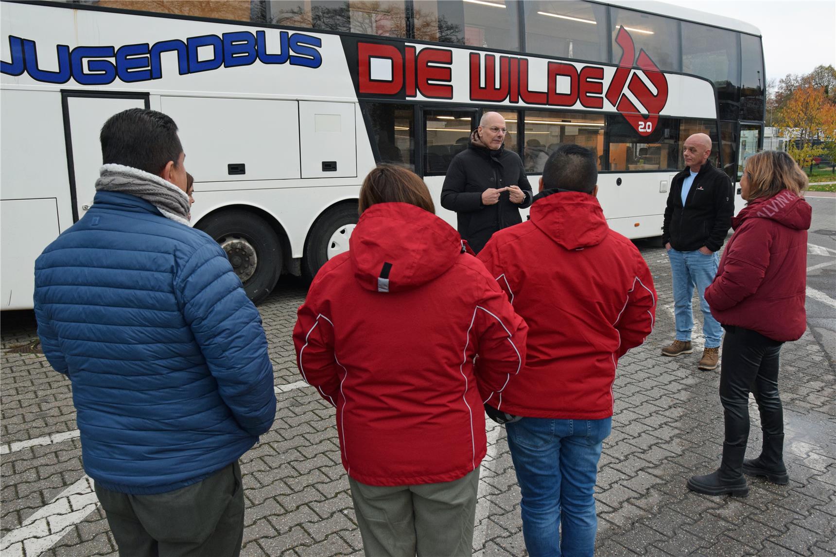 Ein Mann begrüßt eine Menschengruppe, die der Kamera den Rücken zuwendet, an einem Bus.