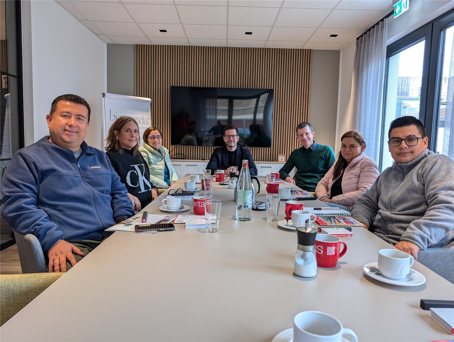 Sieben Personen sitzen in einem Konferenzraum um einen Tisch und schauen in die Kamera