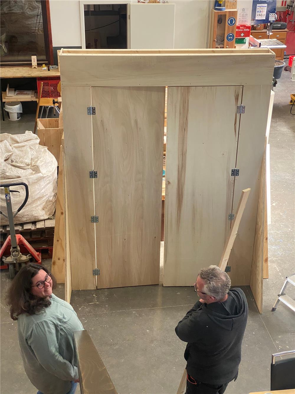 Eine Frau und ein Mann stehen in einer Werkhalle vor einer aus Holz gezimmerten Doppeltür, deren linker Flügel einen Spalt breit offen steht.