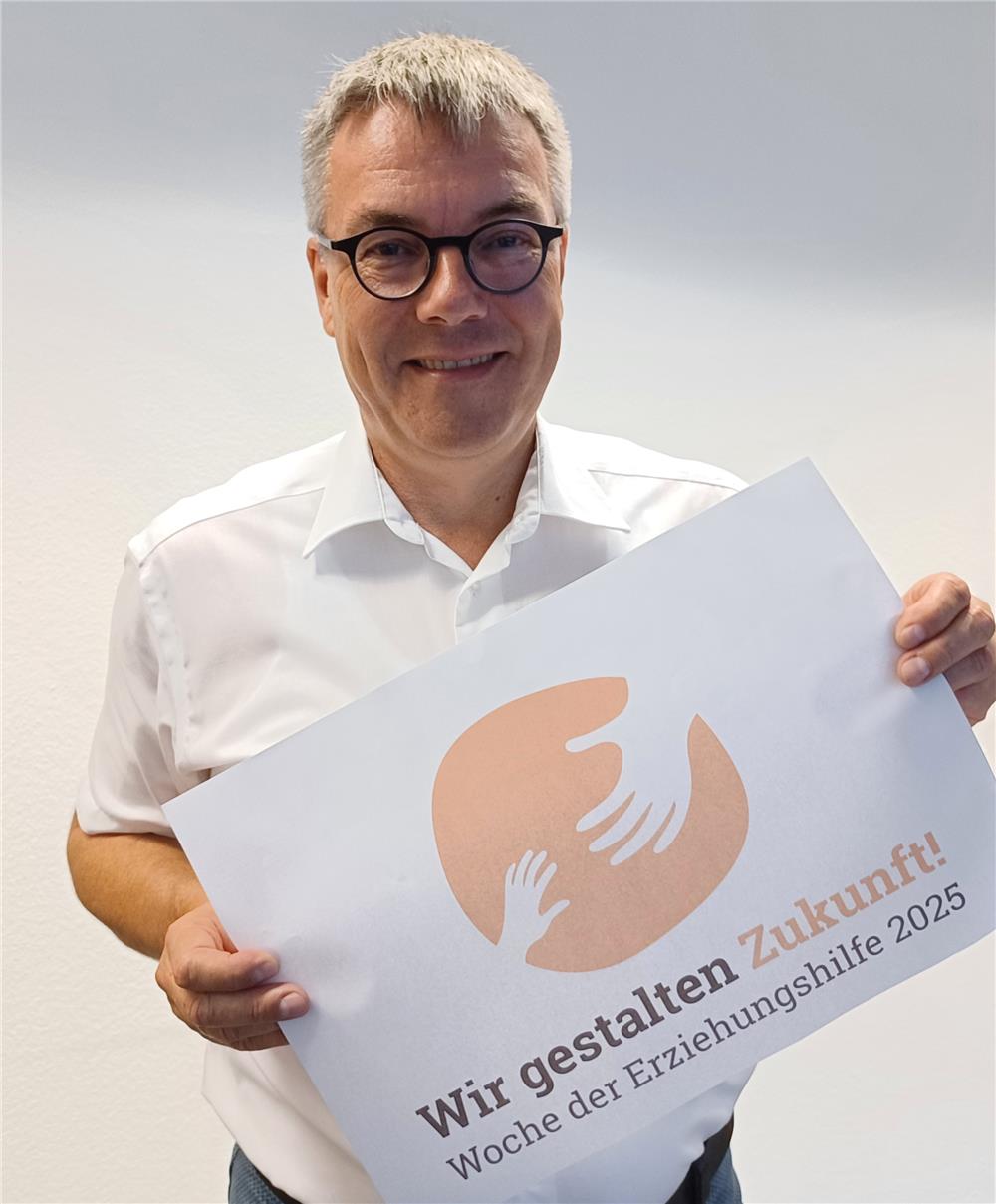 Ein Mann steht vor einem weißen Hintergrund und hält ein weißes Plakat in den Händen. Darauf sind in einem orangen Kreis zwei weiße Hände zu sehen, die sich greifen wollen. Darunter steht der Slogan 