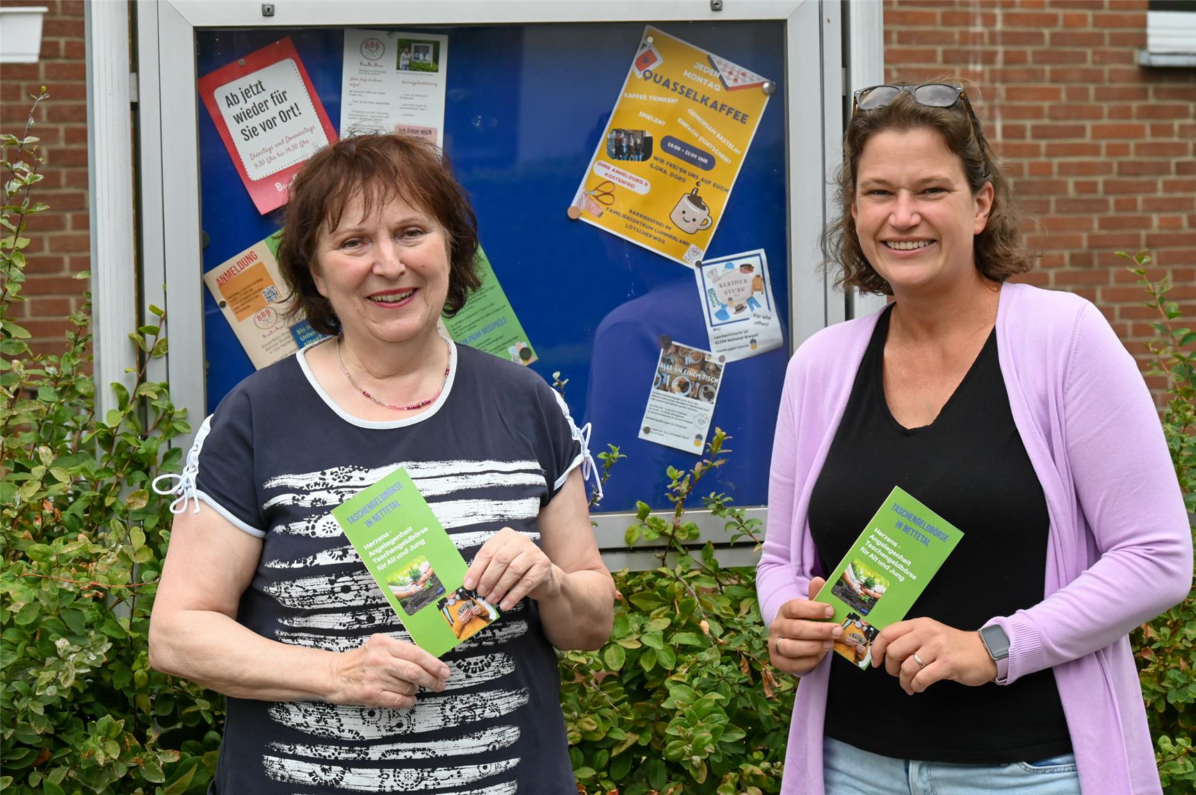 Zwei Frauen stehen links und rechts vor einem Schaukasten mit blauem Hintergrund, der hinter einer grünen Hecke steht. Sie halten grüne Flyer in den Händen.