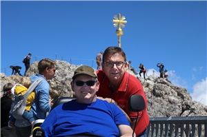 Ein Mann sitzt im Rollstuhl, hinter ihm steht ein Mann. Im Bildhintergrund das Gipfelkreuz der Zugspitze