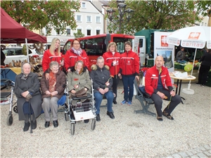 Vier Senioren sitzen am Stadtplatz auf einer Bank. Fünf Pflegekräfte in roten Jacken stehen hinter ihnen und haben jeweils eine Blume in der Hand