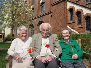Drei Senioren mit Blume sitzen auf einer Bank vor der historischen Fassade des Seniorenheimes.