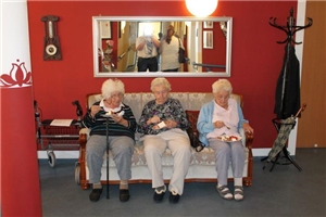 In einem rot gestrichenen Zimmer steht unter einem breiten Spiegel ein altes Sofa. Auf ihm sitzen drei Damen und essen Kuchen.