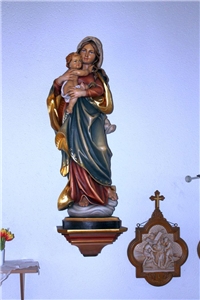 Wir blicken auf eine an der Wand hängenden Marienstatur. Die Heilige Maria hält ihr Kind Jesus mit beiden Händen fest an sich gedrückt. 