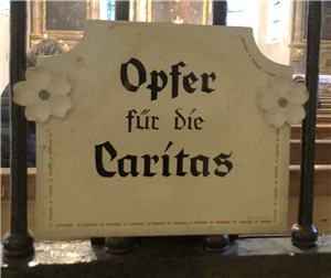 Ein Schild über einem Opferstock. Darauf steht: Opfer für die Caritas!