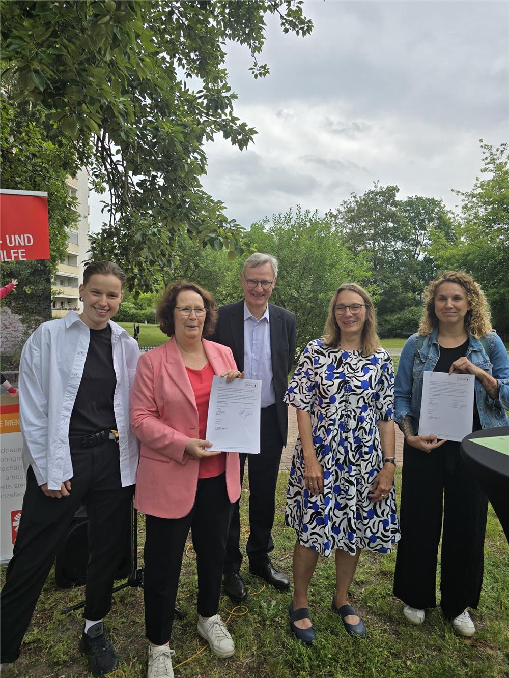 Fünf Personen stehen im Grünen, zwei Frauen halten unterschriebene Dokumente, alle lächeln in die Kamera. Im Hintergrund Bäume, Himmel bewölkt, links ein Caritas-Banner.