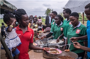Eine afrikanische Dame mit kleinem Kind auf dem Arm hält einen Teller in der Hand und bekommt von Helfenden der Caritas bei der Essensausgabe Lebensmittel serviert. 