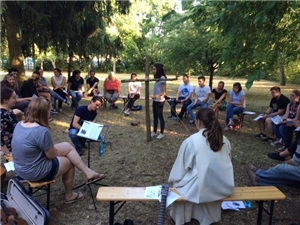Eine Gruppe Menschen sitzt im Garten in einem Kreis um ein Kreuz aus Holz herum.