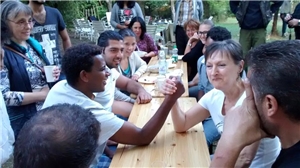 An einem Tisch sitzen verschiedene Menschen unterschiedlicher Nationalitäten zusammen und ein Mann und eine Dame machen "Armdrücken".