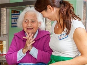 Eine alte Frau lächelt und hält die Hände vor ihrem Oberkörper zusammen, während neben ihr eine junge Dame steht und sich freundlich zu der alten Frau dreht.