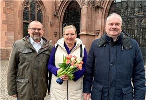 Eine Dame mit Blumen in der Hand steht vor einer Kirche. Rechts und links neben ihr stehen zwei Männer. Alle gucken freundlich in die Kamera.