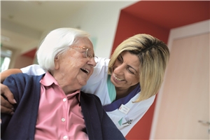 Ein Pflegerin beugt sich zu einer Seniorin herab. Die beiden lächeln sich an.