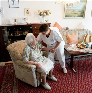 Eine Pflegerin hilft einer Seniorin beim Aufstehen aus ihrem Wohnzimmersessel.