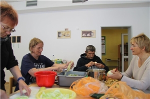 3 Besucher und eine Mitarbeiterin der Tagesstätte sitzen zusammen an einem Tisch und schälen Kartoffeln und Gemüse zur Vorbereitung des Mitagessens.