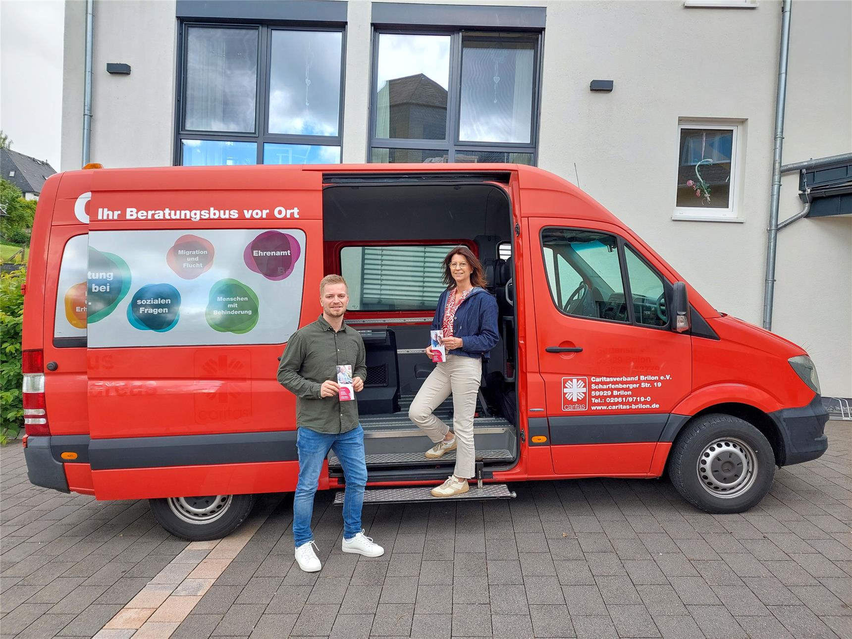 Ein Mann und eine Frau stehen vor einem roten Sprinter und halten Flyer in der Hand. Sie lächeln in die Kamera.