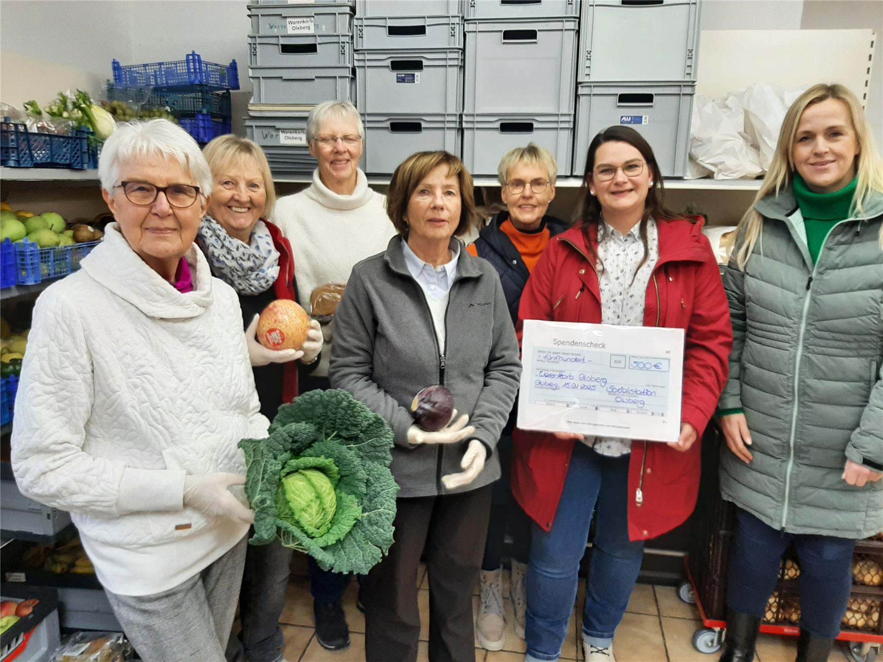 Das Bild zeigt eine Gruppe von sieben Frauen in einem Lagerraum mit Kisten und frischem Obst und Gemüse. Zwei Frauen halten Lebensmittel wie Kohl und Brot, während eine andere eine Spendenurkunde hat.
