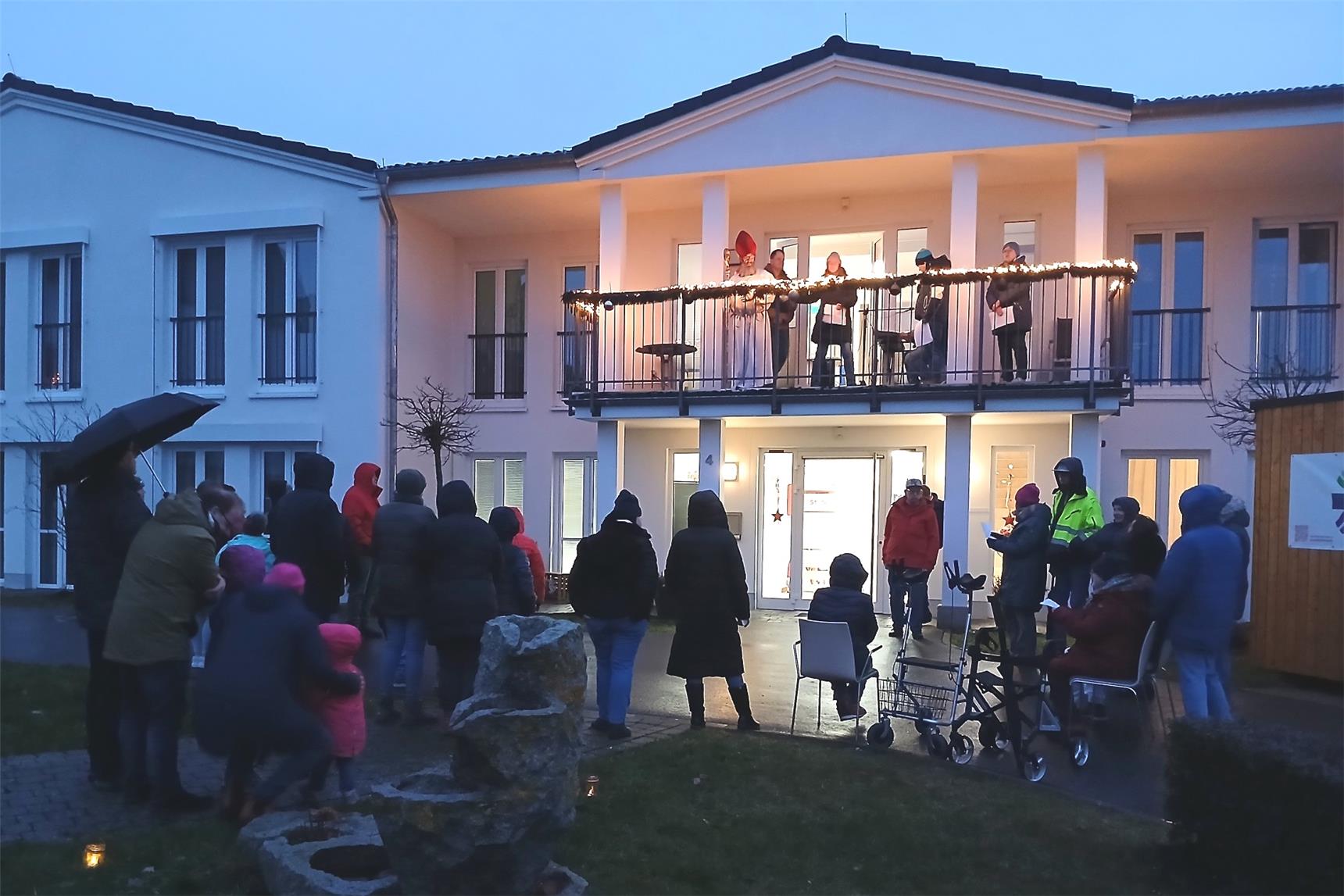 Eine Gruppe Menschen singt vor einem Wohnhaus für Menschen mit Behinderungen Adventlieder. Auf dem Balkon steht eine Person mit Nikolaus-Gewand.