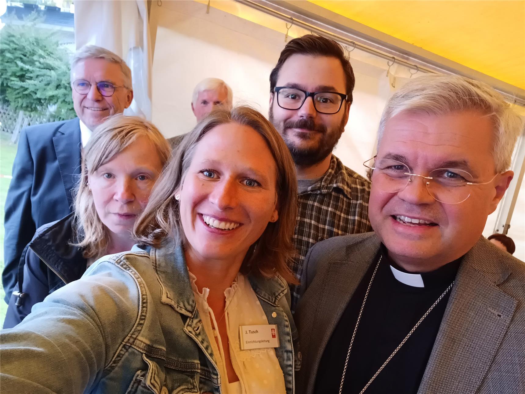 Sieben Menschen stehen zusammen unter einem gelben Zelt. Eine Frau mit Jeansjacke macht ein Selfie. Sie lächelt und hält ein Handy. Neben ihr steht ein Mann mit dunkler Kleidung und einer Kreuz-Kette. Das ist Erzbischof Dr. Udo Markus Bentz. Die anderen Menschen stehen dicht beieinander und schauen freundlich in die Kamera. Im Hintergrund sieht man grüne Pflanzen. Die Stimmung ist fröhlich.
