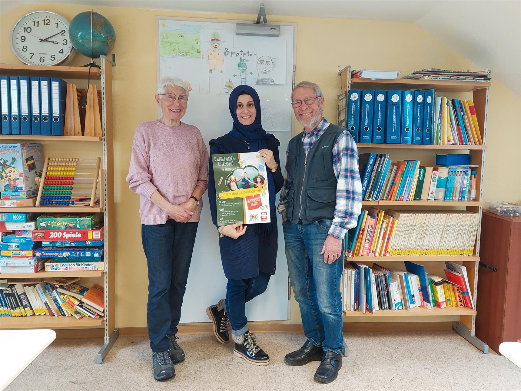 Drei Personen in einem Lernraum, die ein Plakat zur Hausaufgabenbetreuung halten und lächeln.