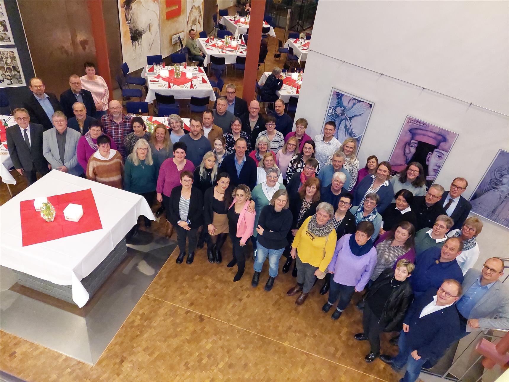 Eine große Gruppe von Menschen steht in einem festlich dekorierten Raum und blickt lächelnd zur Kamera. Im Hintergrund sind gedeckte Tische mit roten Tischdecken und Kerzen sowie Kunstwerke.