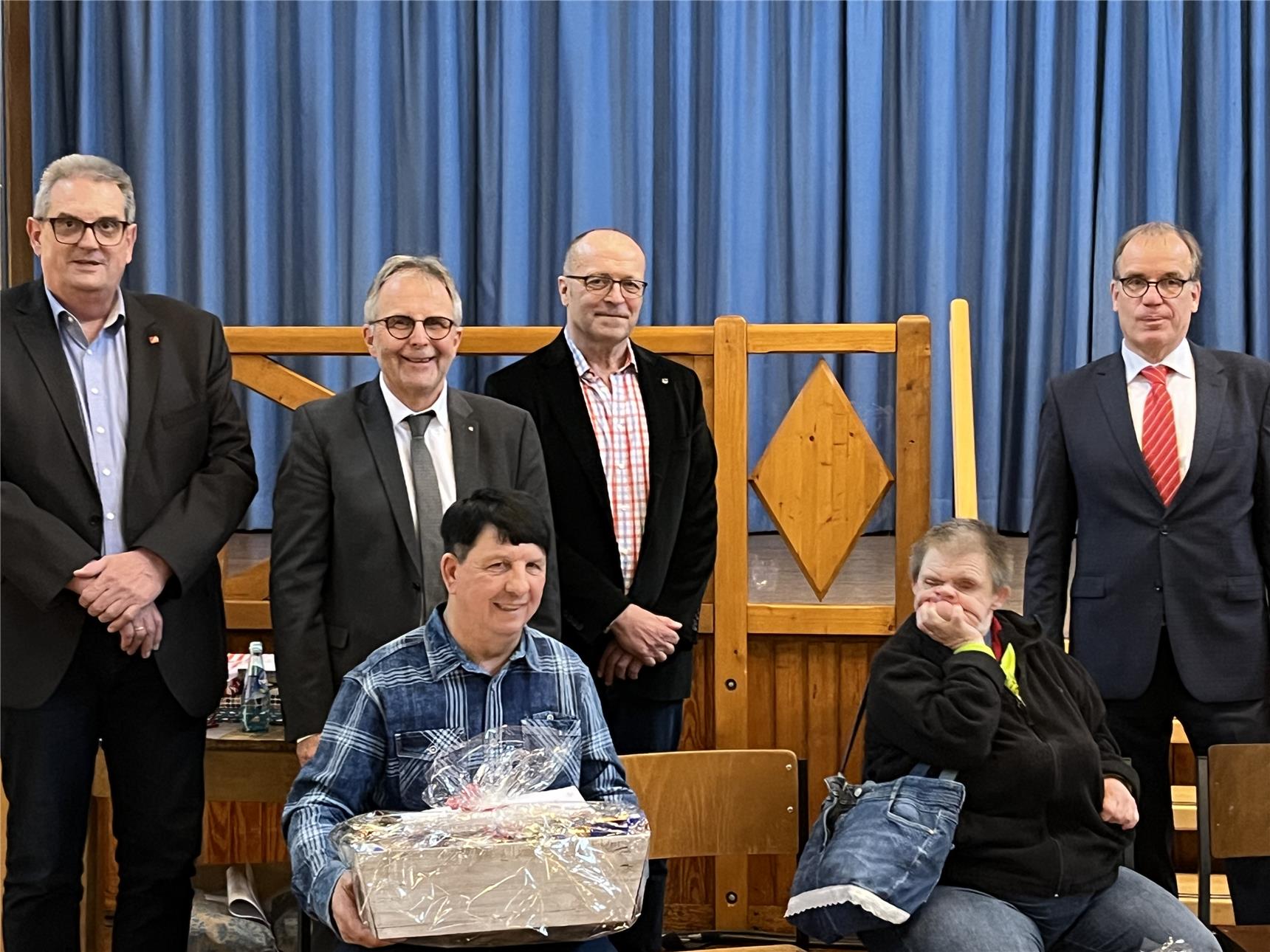 Dieses Bild zeigt eine Gruppe von sechs Personen, davon vier stehend im Hintergrund und zwei sitzend im Vordergrund. Eine der sitzenden Personen hält einen Geschenkkorb, der in Zellophan verpackt ist,