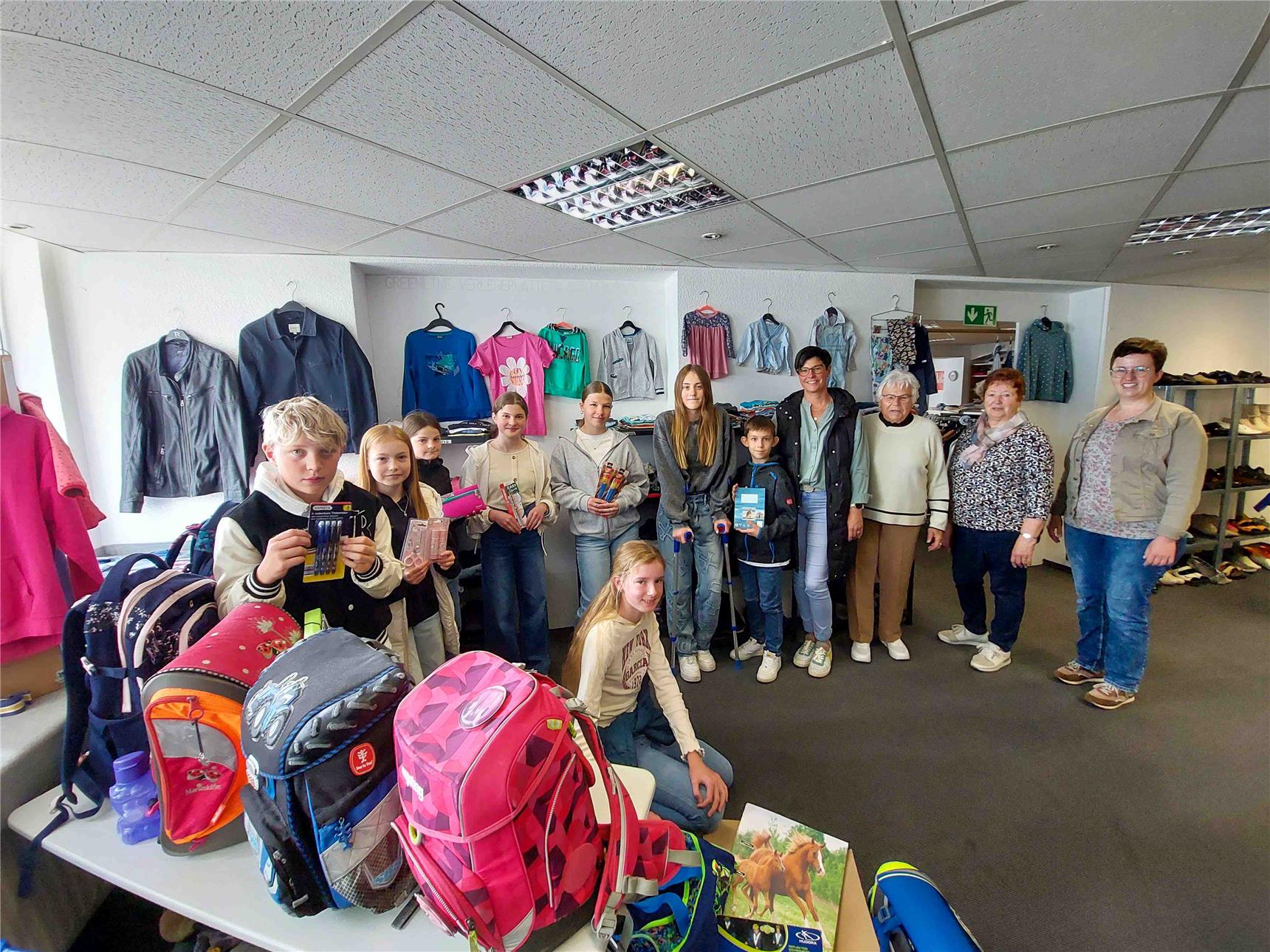 Eine Gruppe von Kindern und Erwachsenen steht gemeinsam in einem Raum mit Kleider- und Schuhausgabe. Die Kinder halten Schulmaterialien wie Stifte, Hefte und Federmäppchen in der Hand, vor ihnen stehen bunte Schulrucksäcke auf einem Tisch. Im Hintergrund hängen Kleidung und Jacken an der Wand. Es geht um die Ausgabeaktion für die Aktion Smarter Schulstart.