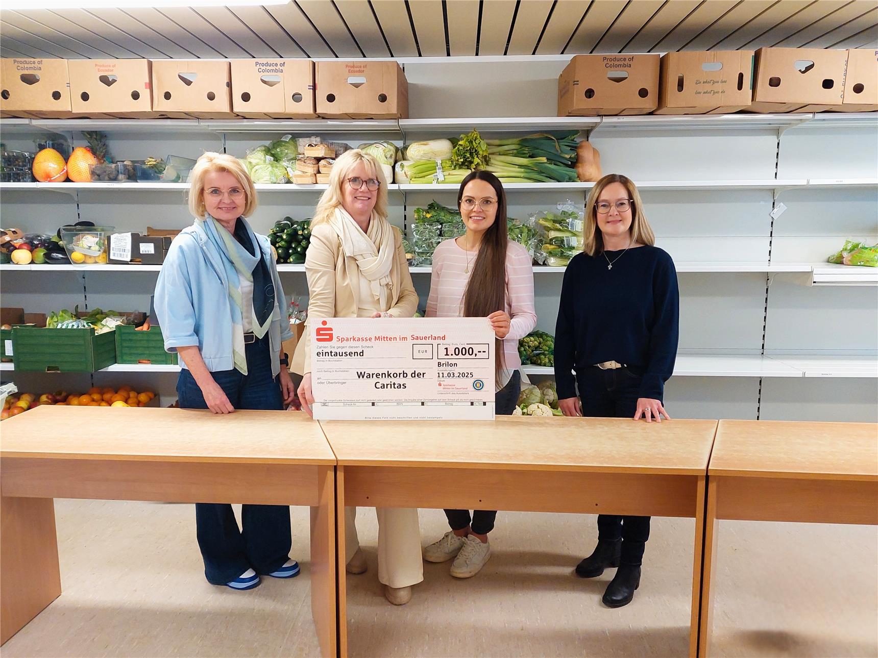 Vier Frauen stehen im Warenkorb vor Regalen mit frischem Obst und Gemüse. Zwei von ihnen halten einen großen symbolischen Scheck über 1.000 Euro von der Sparkasse.
