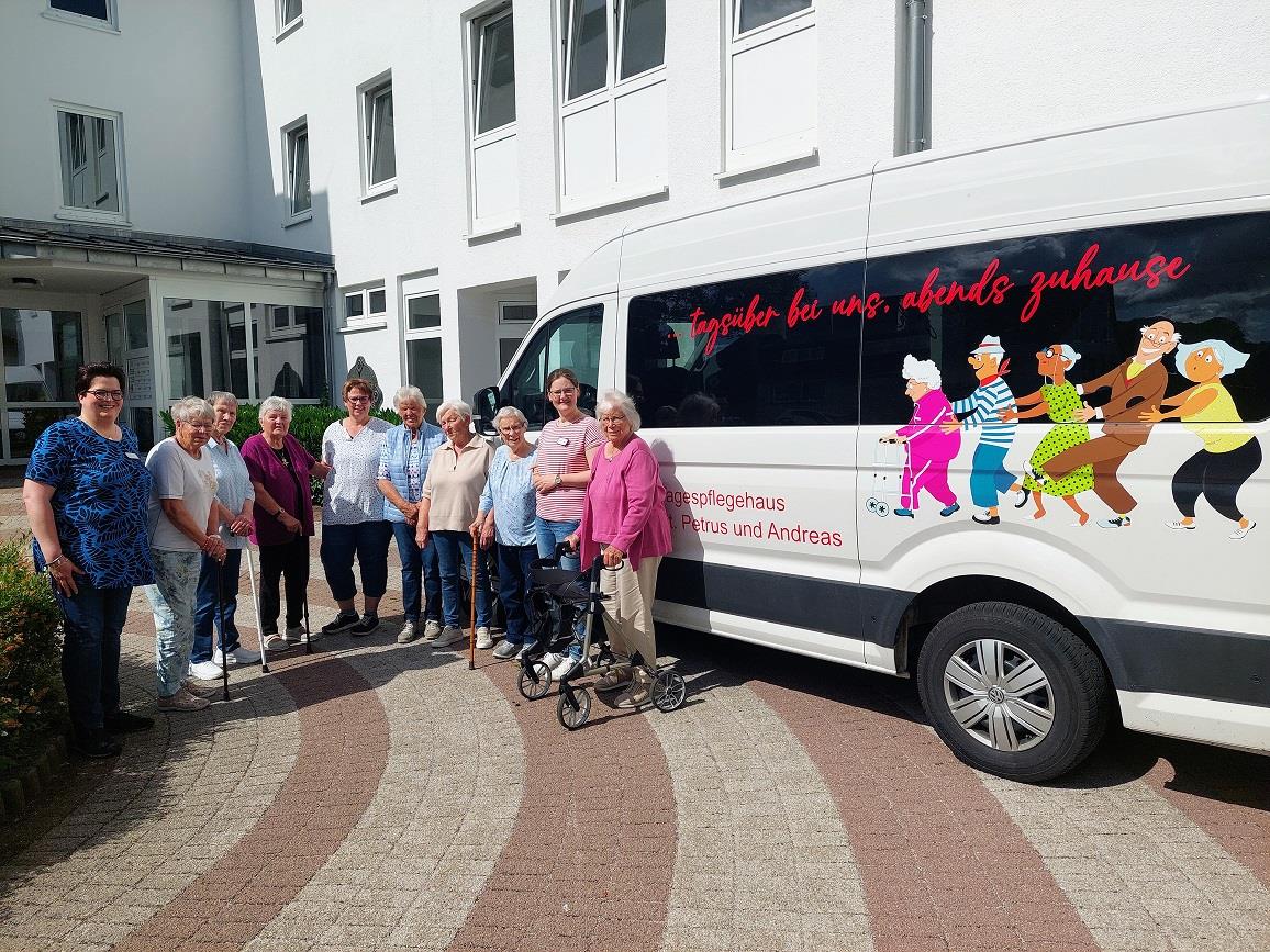 Eine Gruppe Frauen steht vor einem Kleinbus mit der Aufschrift: Tagespflegehaus St. Petrus und Andreas. Die Atmosphäre ist heiter. Alle Frauen lachen in die Kamera. 