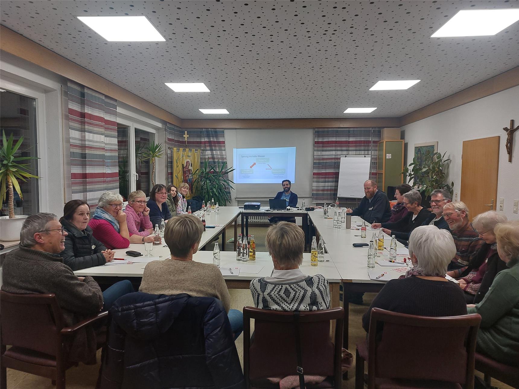 Ein Raum mit rechteckiger Tischanordnung, an der 16 Personen unterschiedlichen Alters sitzen. Sie hören einem Vortragenden zu, der am Kopfende vor einem Beamer sitzt, auf dem eine Präsentationsfolie z