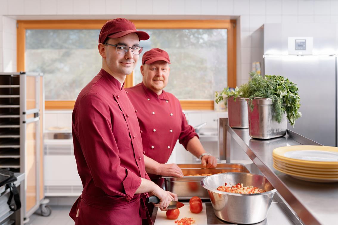 Junger und Mann mittleren Alters in roter Kochkleidung kochen gerade und lächeln Richtung Kamera