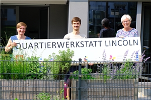 Zwei Frauen und ein Mann vor einem Gebäude halten ein Schild mit der Aufschrift Quartierwerkstatt Lincoln.