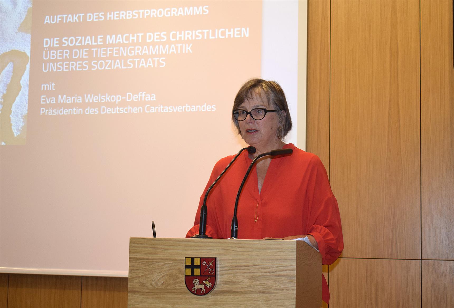 Eine Frau im roten Kleid steht an einem Podium und hält einen Vortrag.