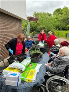Senior:innen und Pflegerinnen sitzen im Freien an einem Tisch und bepflanzen Balkonkästen