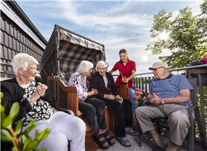 Senior:innen sitzen bei strahlendem Sonnenschein gemeinsam auf einer Terrasse, zwei davon in einem Starndkorb. Eine Pflegekraft reicht ein Getränk.