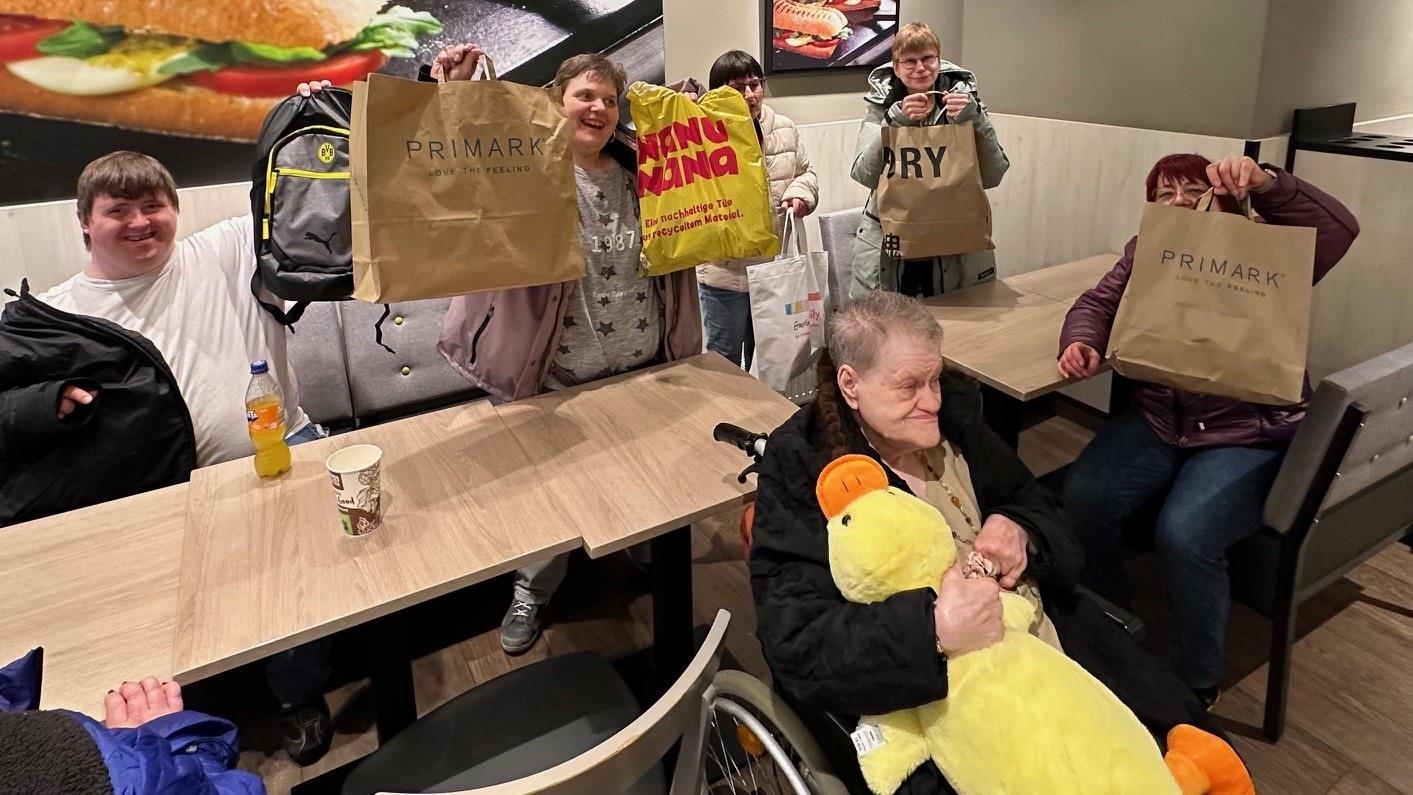 Menschen mit Beeinträchtigung sitzen in einem Café und halten Einkaufstüten in die Höhe.