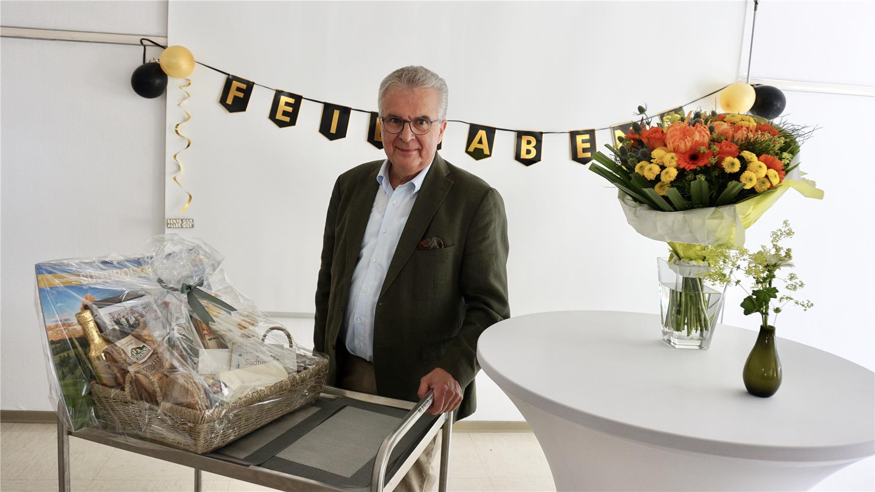 Rolf Mull lächelt in die Kamera, rechts im Bild ein Stehtisch mit Blumenstrauss, links im Bild der große Präsentkorb