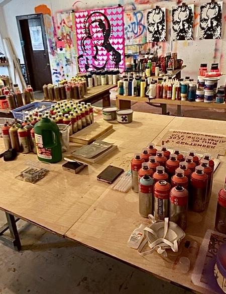Im Atelier: Man sieht im vorderen Bereich auf mehreren Werktischen zahlreiche Farbdosen und Utensilien, hinten an der Wand hängen Kunstwerke