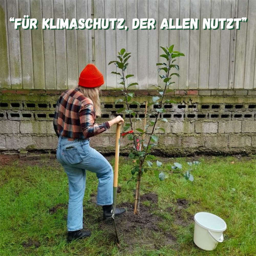 Junge Frau steht mit Schaufel und Eimer neben einem kleinen Baum, den sie gerade gepflanzt hat