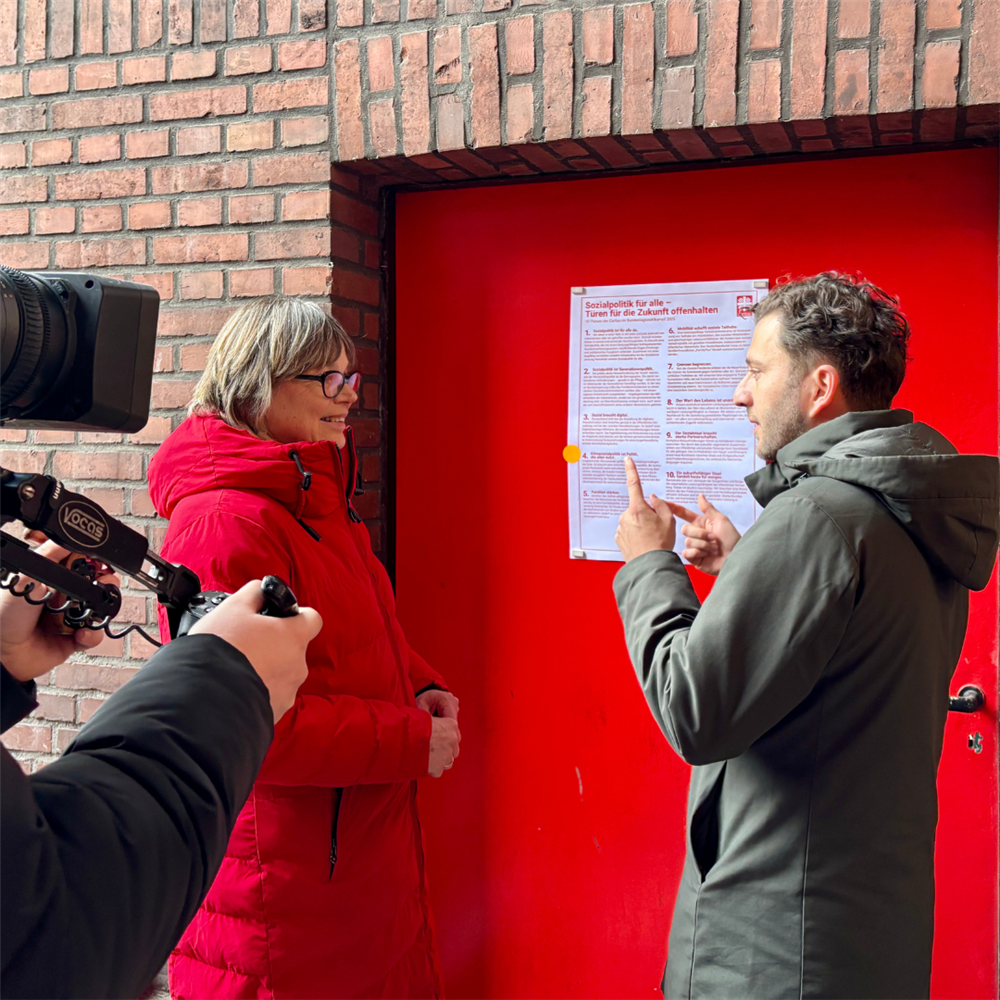 Eine Kamera filmt Welskop-Deffaa und Banaszak vor einer roten Türe mit Thesenplakat
