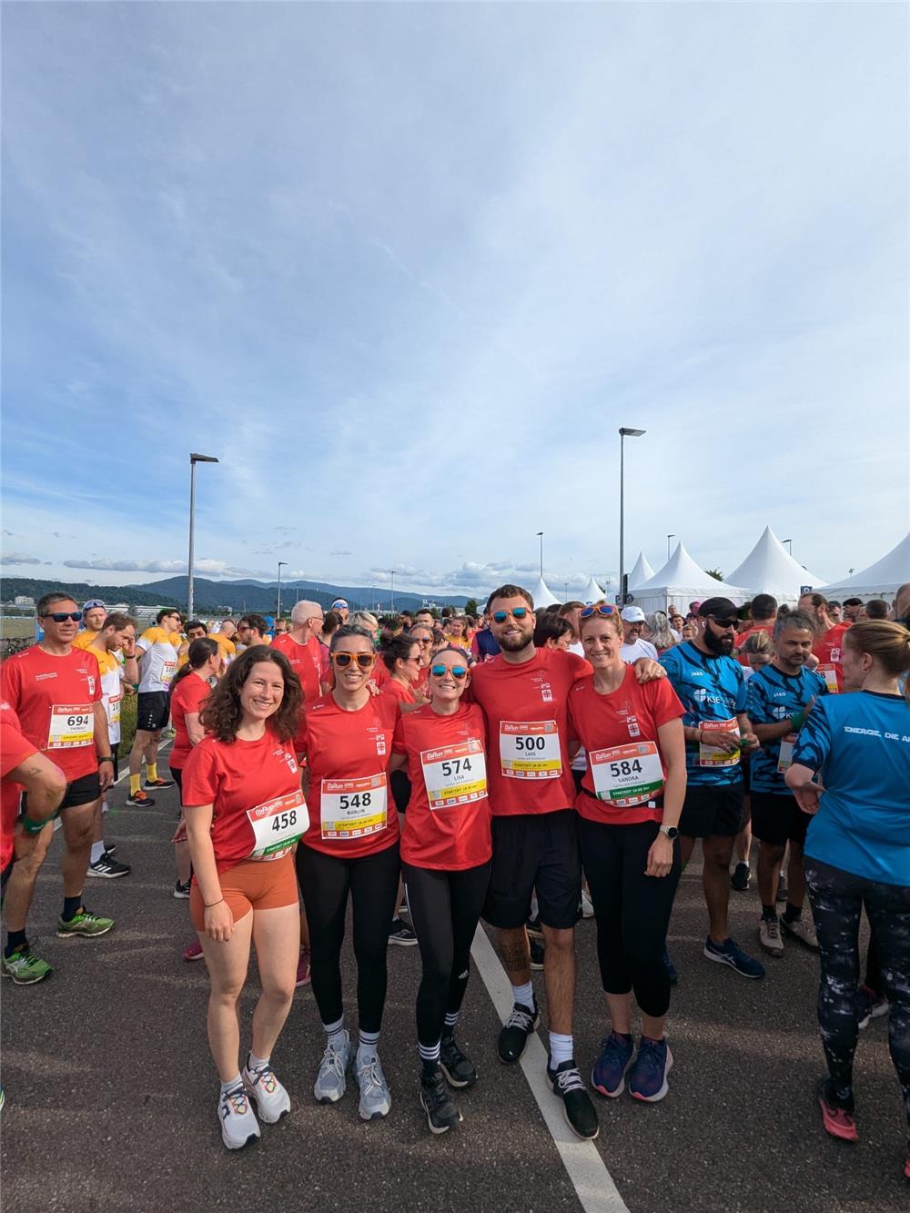 Das Bild zeigt viele Teilnehmende des Firmenlaufes. Im Vordergrund die fünf des Caritasverbandes für den Landkreis Emmendingen. Im Hintergrund sieht man die Zelte, die der Veranstalter für den Lauf aufgebaut hat. Der Himmel ist fast wolkenlos. 