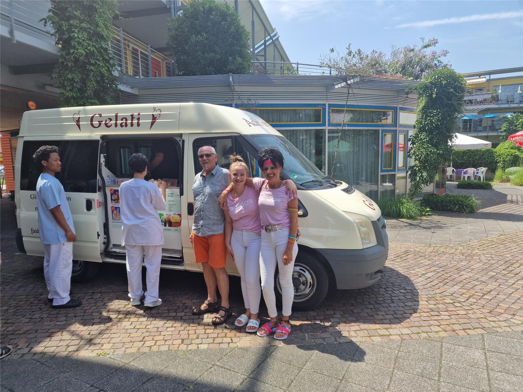 Das Bild zeigt einen Eiswagen, fünf Menschen stehen davor, einer verteilt das Eis und die anderen freuen sich über die schöne Erfrischung. Der Eiswagen steht im Innenhof des Seniorenzentrums, im Hintergrund das Café Plausch, der Veranstaltungsraum.