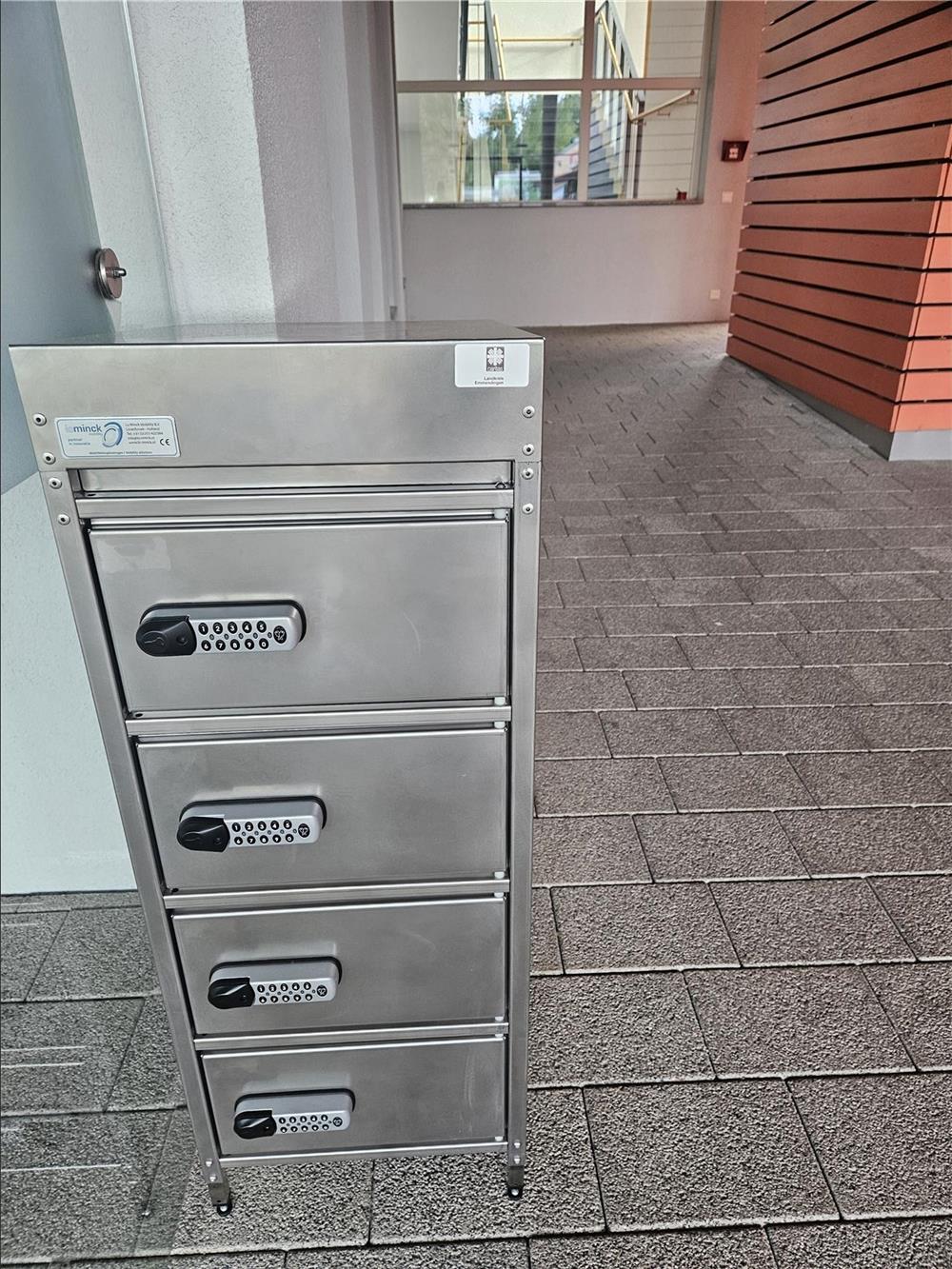 Das Bild zeigt einen Metallschrank mit vier Fächern, in die ein Akku gelegt werden kann. Seitlich ist der Zwischenraum zwischen der Verwaltung und der Seniorenwohnanlage zu sehen. 