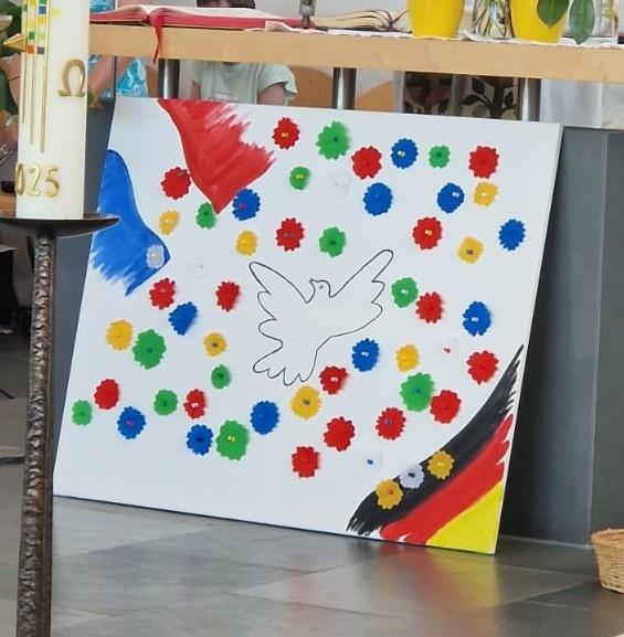 Das Bild zeigt ein Bild mit einer Friedenstaube und den Flaggen von Deutschland und Frankreich. Dieses Bild steht vor dem Altar und am linken Rand ist ein Teil der Osterkerze zu erkennen. 