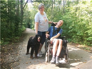 Eine Frau, ein junger Mann im Rollstuhl und ein Hund auf einem Waldweg