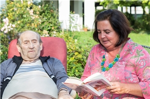 Ein Gast sitzt mit einer Ehrenamtlichen im Garten; sie liest ihm aus einem Buch vor.