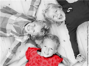 3 Kindergartenkinder liegen am Rücken auf d. Boden, Köpfe zueinander, sich an d. Händen haltend, lachen in Kamera. In Graustufen, 1 Shirt rot. © contrastwerkstatt|stock.adobe.com|bearb.Caritas FRG