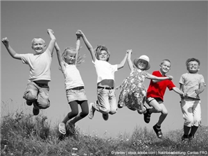 6 Kinder springen auf einer Sommerwiese in d. Luft. Lachen ausgelassen in die Kamera. In Graustufen. Vorletzte Kind (re.) hat ein rotes Shirt. © yanlev|stock.adobe.com|bearb.Caritas FRG